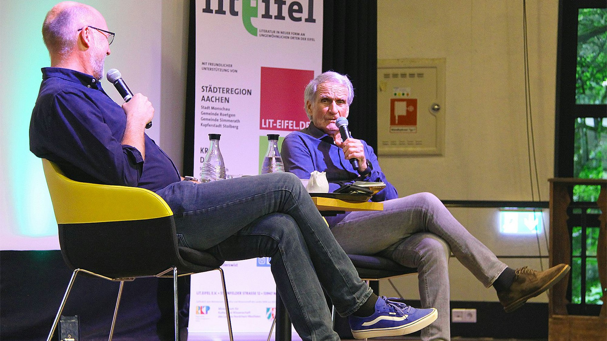 Sven Pistor und Wolfgang Overath sitzen auf einer Bühne und unterhalten sich.