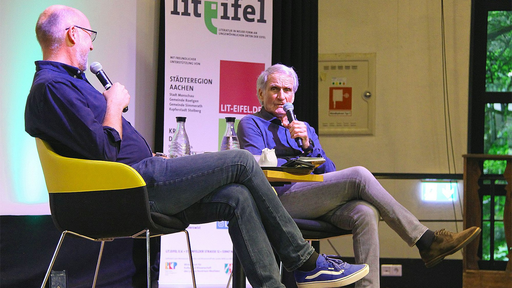 Zwei Männer sitzen auf einer Bühne und unterhalten sich.