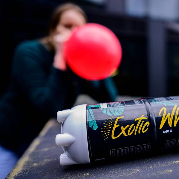 Eine Flasche Lachgas, im Hintergrund hat eine junge Person einen vollen Luftballon am Mund