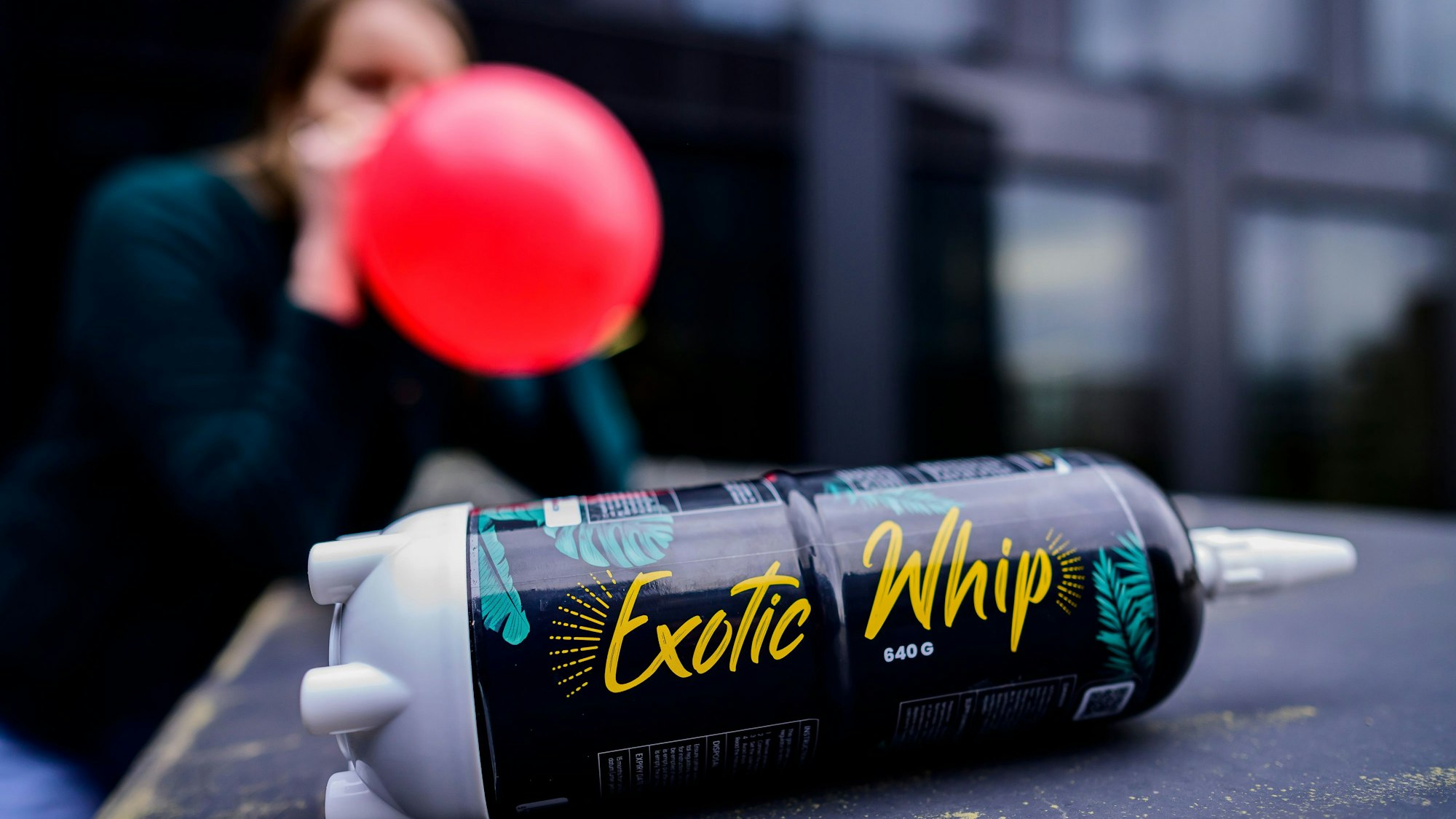 Eine Flasche Lachgas, im Hintergrund hat eine junge Person einen vollen Luftballon am Mund
