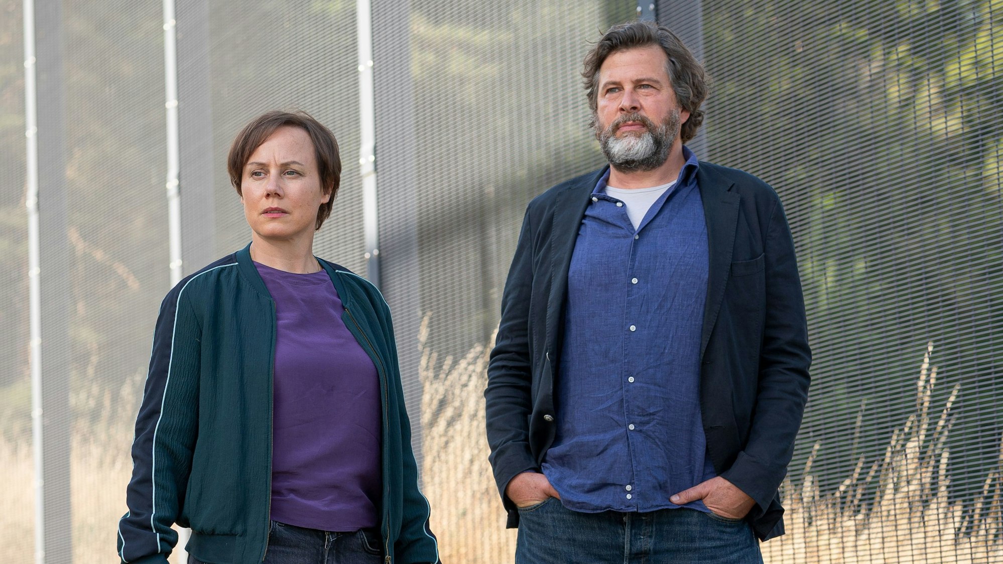 Auf dem Bild stehen die beiden Ermittler vor dem Schutzzaun der forensischen Klinik.