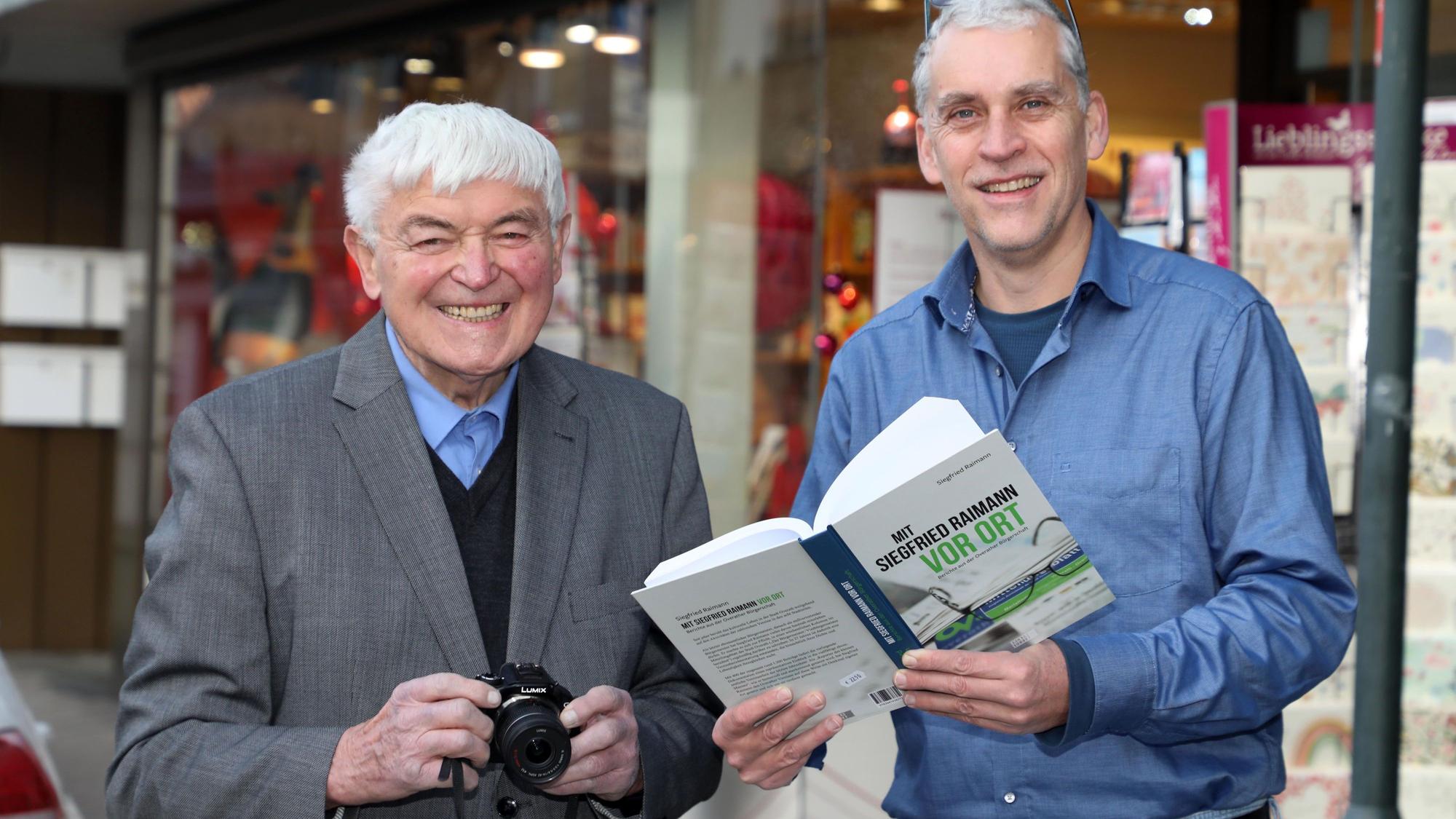 Siegfried Raimann steht mit einer Kamera in der Hand neben Verleger und Buchhändler Alexander Bücken, der sein Buch „Mit Siegfried Raimann vor Ort“ in den Händen hält.