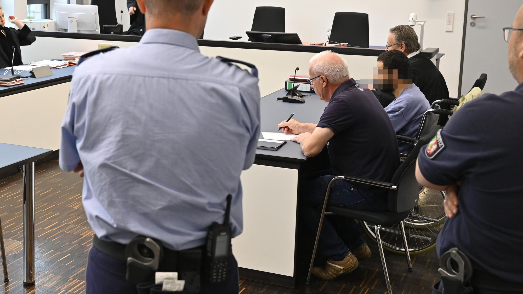 Der Angeklagte sitzt in einem Gerichtssaal neben seinem Verteidiger in einem Rollstuhl.