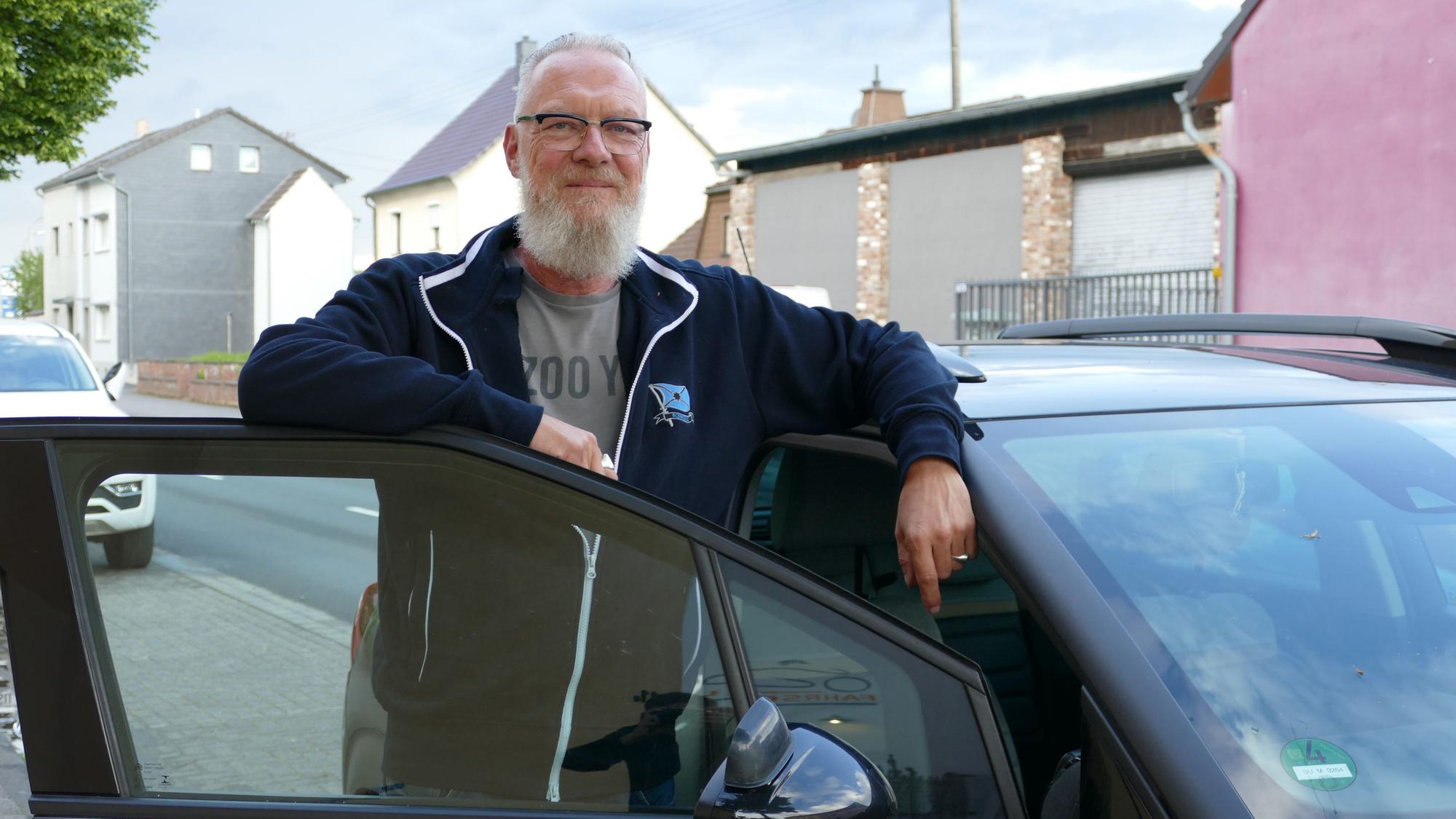 Ein Mann mit weißem Bart und weißen Haaren, blauer Jacke und Brille neben einem Auto. Er lehnt sich auf die geöffnete Tür.