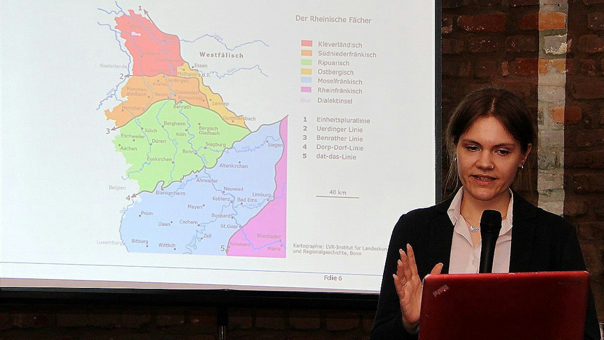 Sprachwissenschaftlerin Verena Krautwald hält einen Vortrag. Hinter ihr wird über einen Beamer eine Skizze der Dialektgebiete im Rheinland an die Wand projiziert.