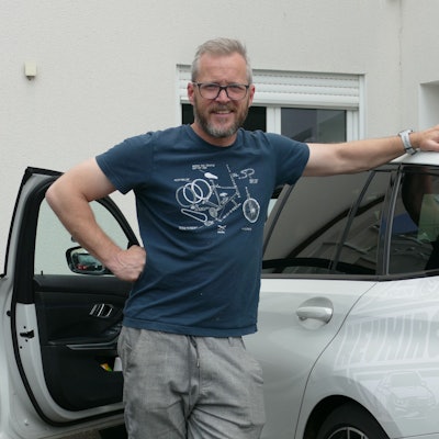 Ein Mann im blauen T-Shirt steht in der geöffneten Tür neben einem weißen Kombi.