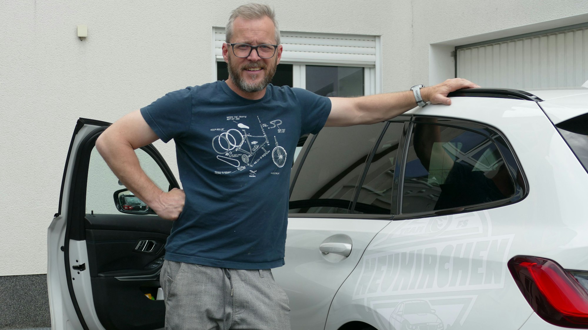 Ein Mann im blauen T-Shirt steht in der geöffneten Tür neben einem weißen Kombi.