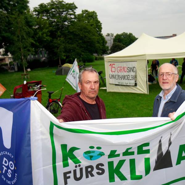 Walter Harings und Christoph Schulenkorf (v.r.) stehen hinter eine Flagge auf der „Kölle fürs Klima“ steht. Im Hintergrund ist ein Pavillon zu sehen.