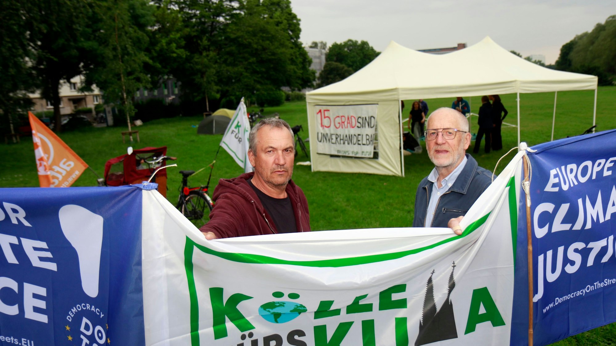 Walter Harings und Christoph Schulenkorf (v.r.) stehen hinter eine Flagge auf der „Kölle fürs Klima“ steht. Im Hintergrund ist ein Pavillon zu sehen.