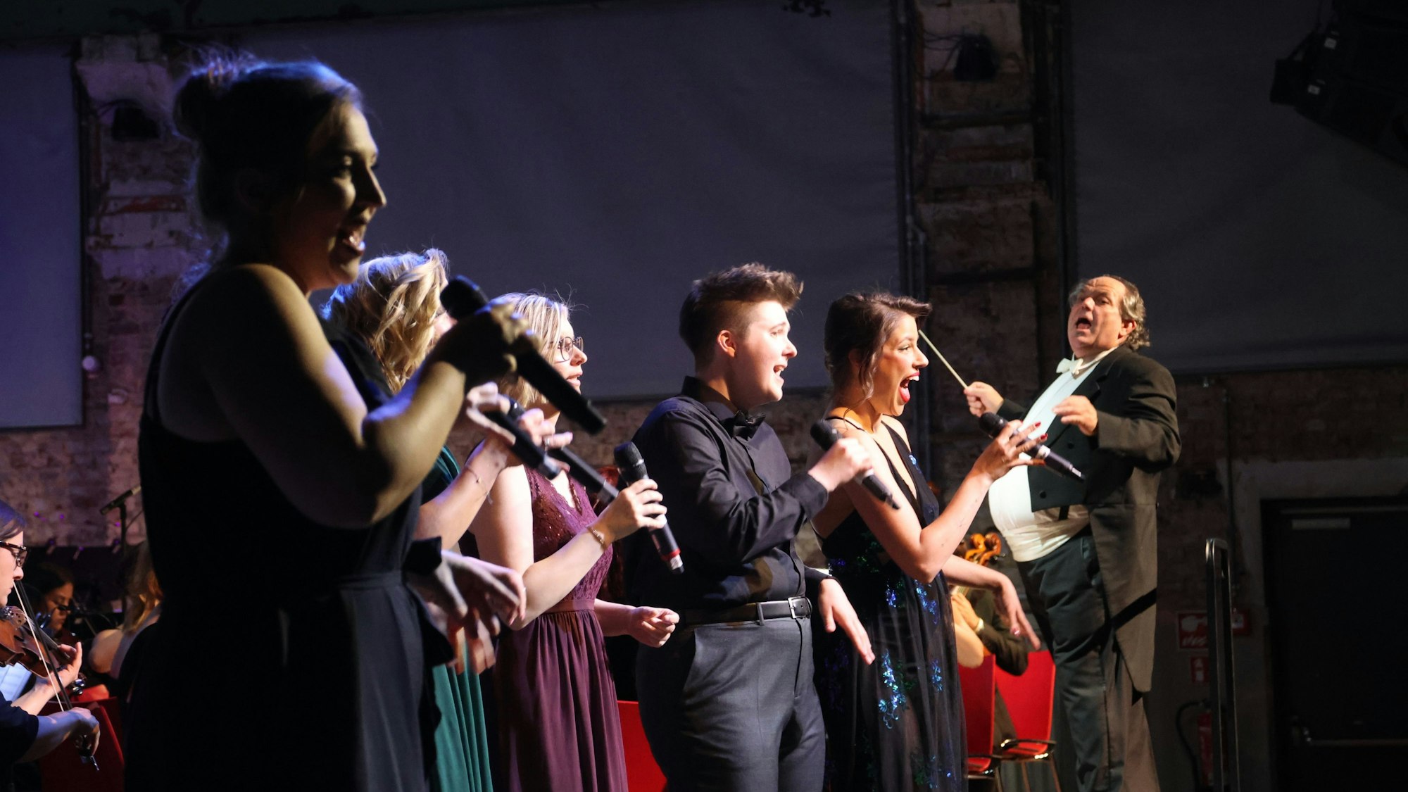 Fünf Sängerinnen und Sänger mit weit geöffneten Mündern singen, im Hintergrund ist ein Dirigent im Frack und mit Taktstock zu sehen.
