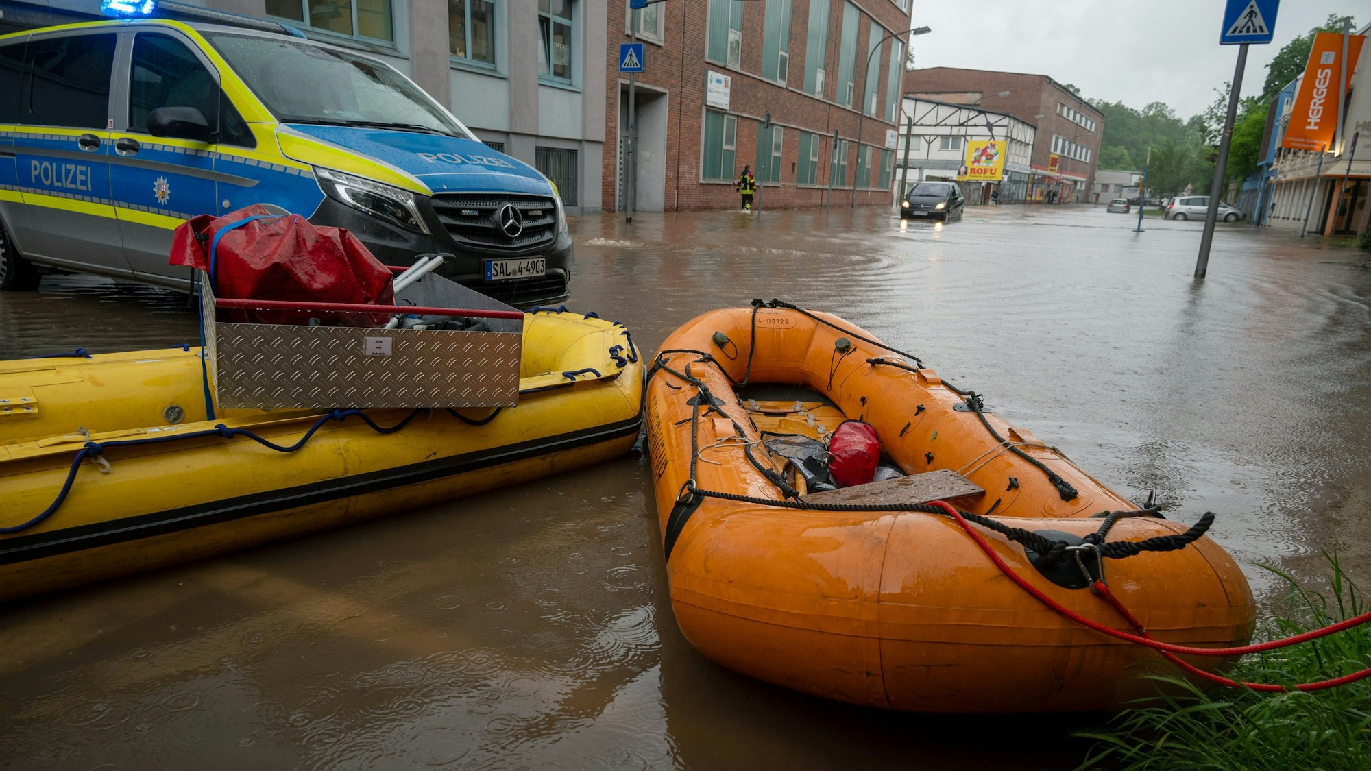 Man sieht eine überschwemmte Straße. Ein Polizeiwagen steht quer. Vor ihm liegen ein gelbes und ein orangenes Schlauchboot bereit.