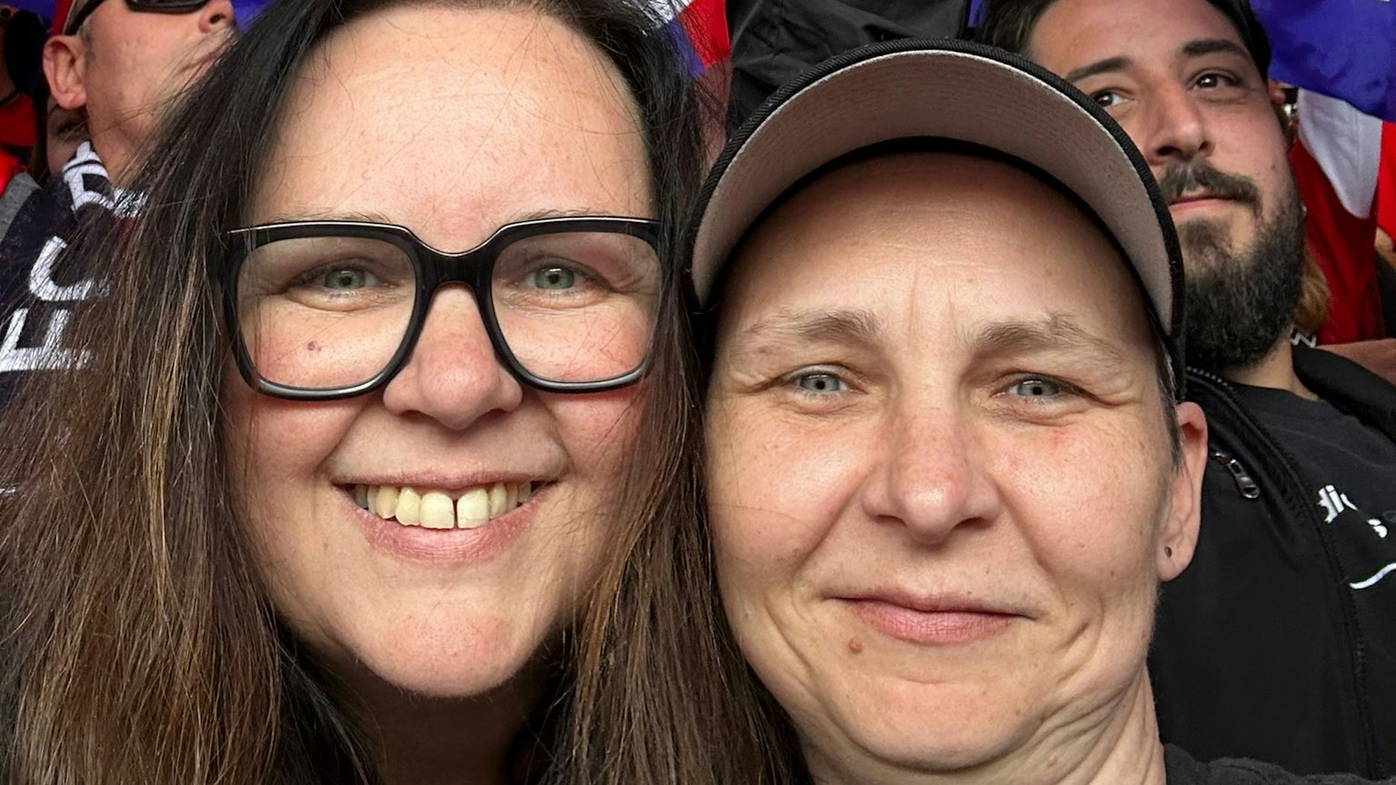 Zwei Frauen lächeln in die Kamera. Hinter ihnen schwenken Fußball-Fans Fahnen.