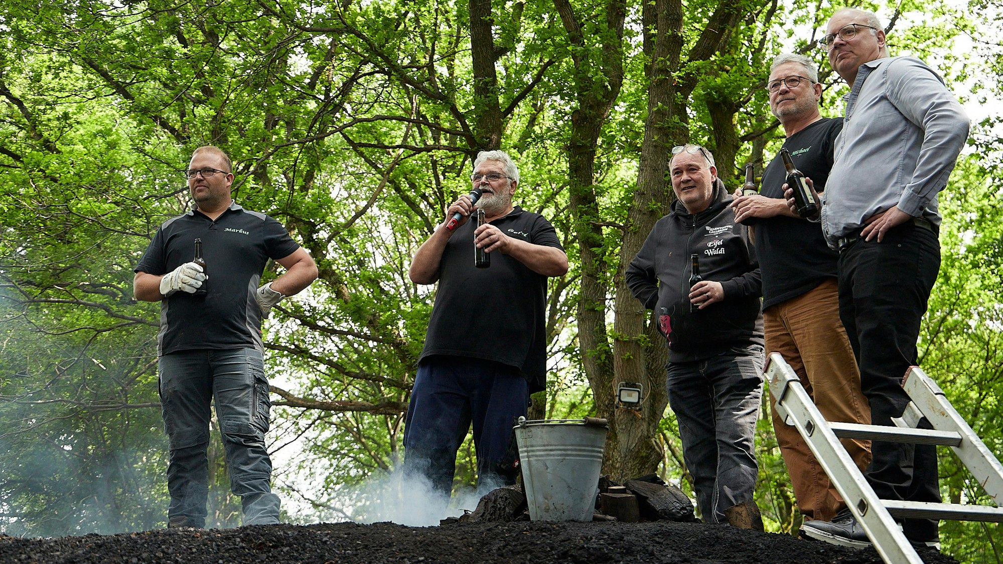 Markus Linden, Gerd Linden, Walter Lehnertz, Michael Lammertz und Jochen Weiler stehen an dem qualmenden Kohlemeiler und halten Bierflaschen in der Hand.
