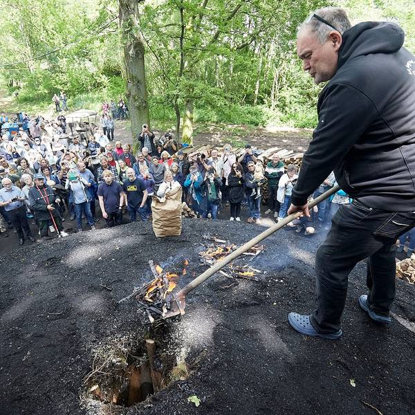 Walter Lehnertz steht auf einem schwarzen Hügel und hält eine Schaufel mit Glut in der Hand. Rund um den Kohlemeiler stehen zahlreiche Zuschauerinnen und Zuschauer.