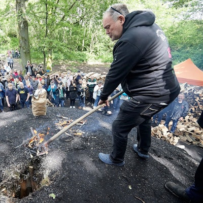 Walter Lehnertz steht auf einem schwarzen Hügel und hält eine Schaufel mit Glut in der Hand. Rund um den Kohlemeiler stehen zahlreiche Zuschauerinnen und Zuschauer.