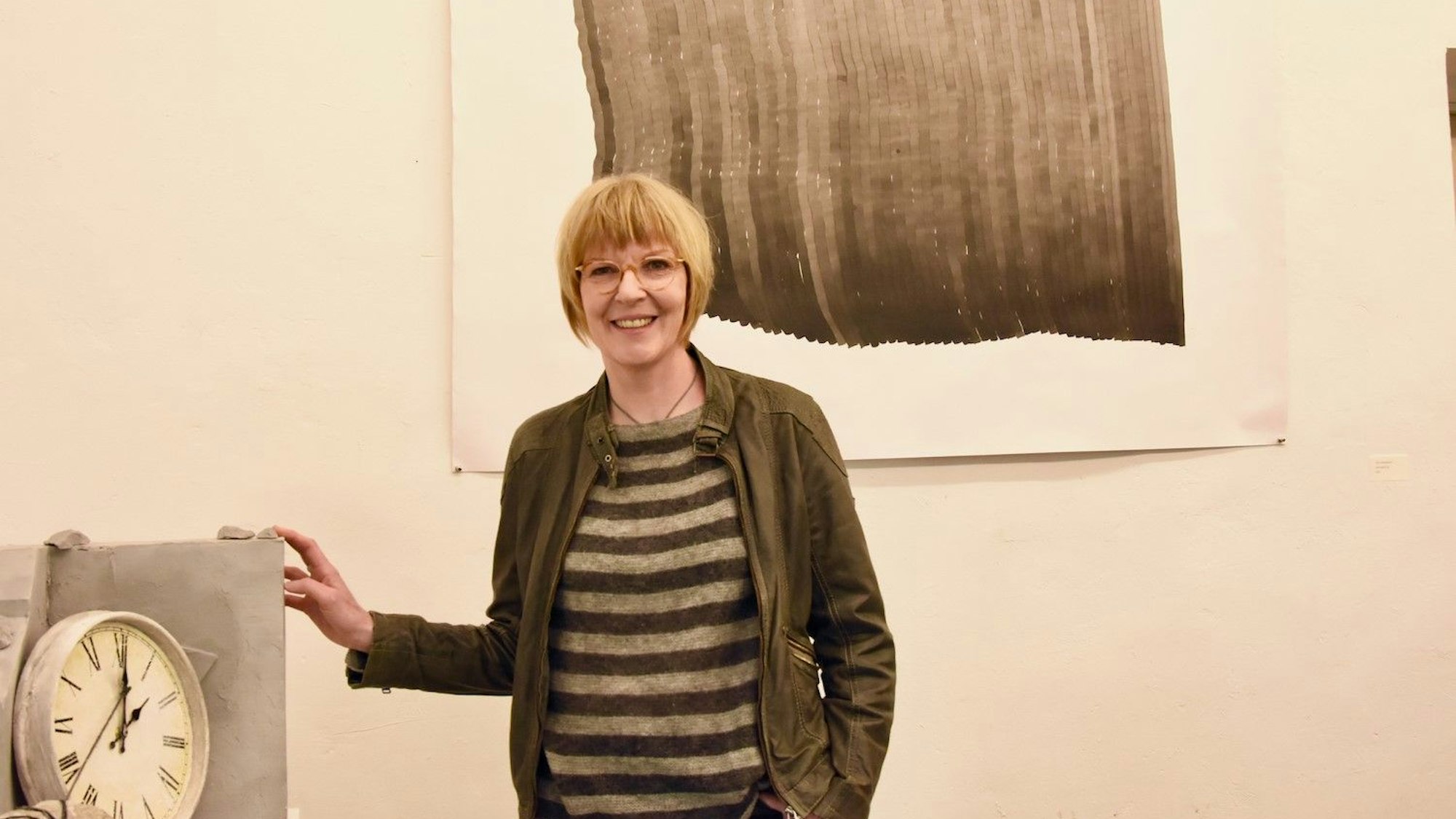 Eine Frau mit Pagenkopf-Frisur steht vor einem Gemälde, das an der Wand hängt und neben einer Wanduhr.