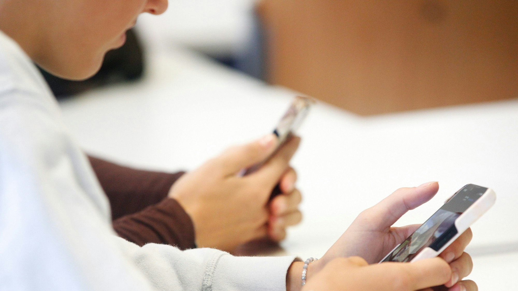 Man sieht zwei Schüler, die in einem Klassenraum nebeneinander sitzen und beide ihre Smartphones in den Händen halten und darauf gucken.