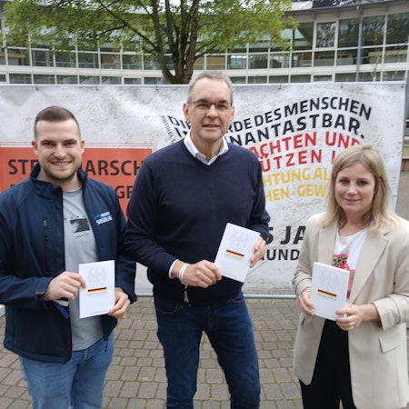 Zwei Männer und eine Frau mit Büchern in der Hand vor einem Plakat