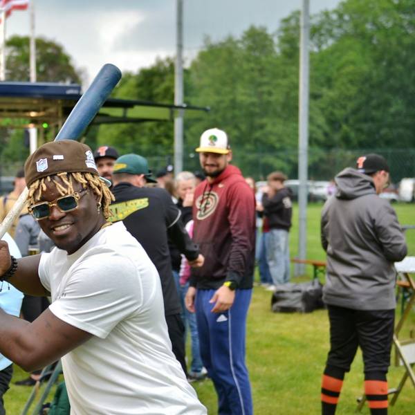 Ein Baseballspieler mit schwarzer Hautfarbe und blonden Dreadlocks hält einen Baseballschläger und lacht in die Kamera. Im Hintergrund sind weitere Baseballspieler zu sehen.