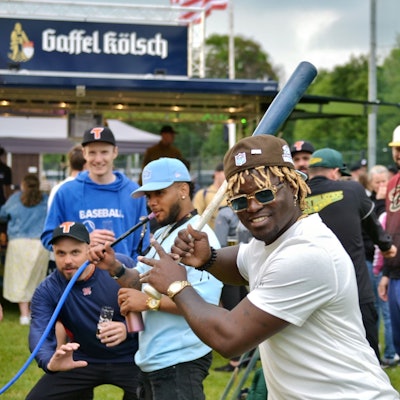 Ein Baseballspieler mit schwarzer Hautfarbe und blonden Dreadlocks hält einen Baseballschläger und lacht in die Kamera. Im Hintergrund sind weitere Baseballspieler zu sehen.