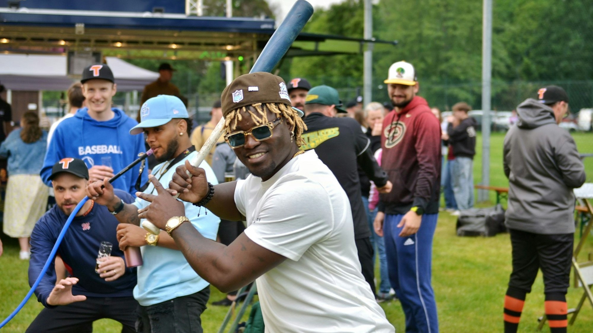 Ein Baseballspieler mit schwarzer Hautfarbe und blonden Dreadlocks hält einen Baseballschläger und lacht in die Kamera. Im Hintergrund sind weitere Baseballspieler zu sehen.