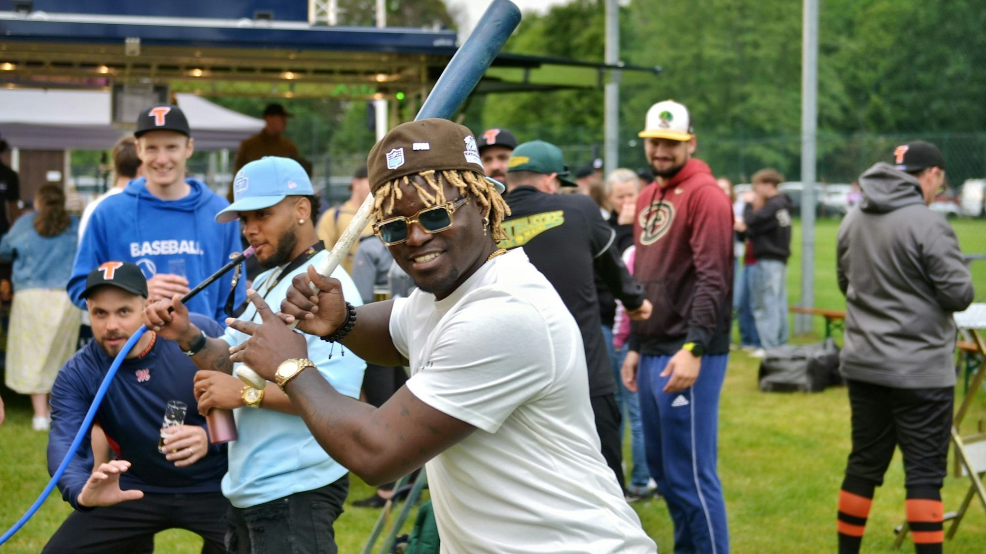 Ein Spieler mit blonden Rastalocken unter einer Kappe hält einen Baseballschläger in den Händen, im Hintergrund zieht jemand am Schlauch einer Bong.