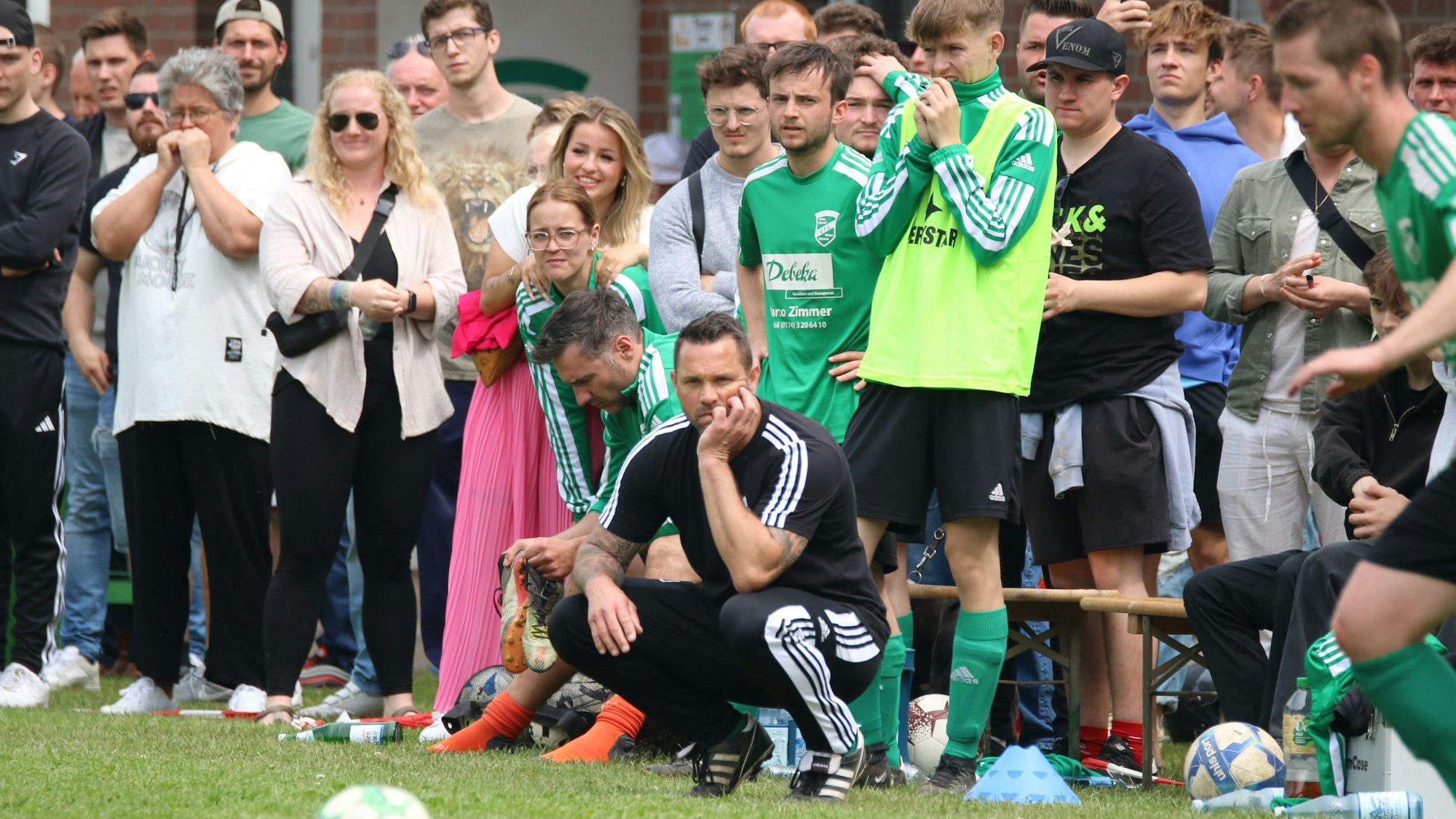 Ein Fußballtrainer verfolgt hockend das Geschehen, hinter ihm Ersatzspieler und viele Zuschauer.