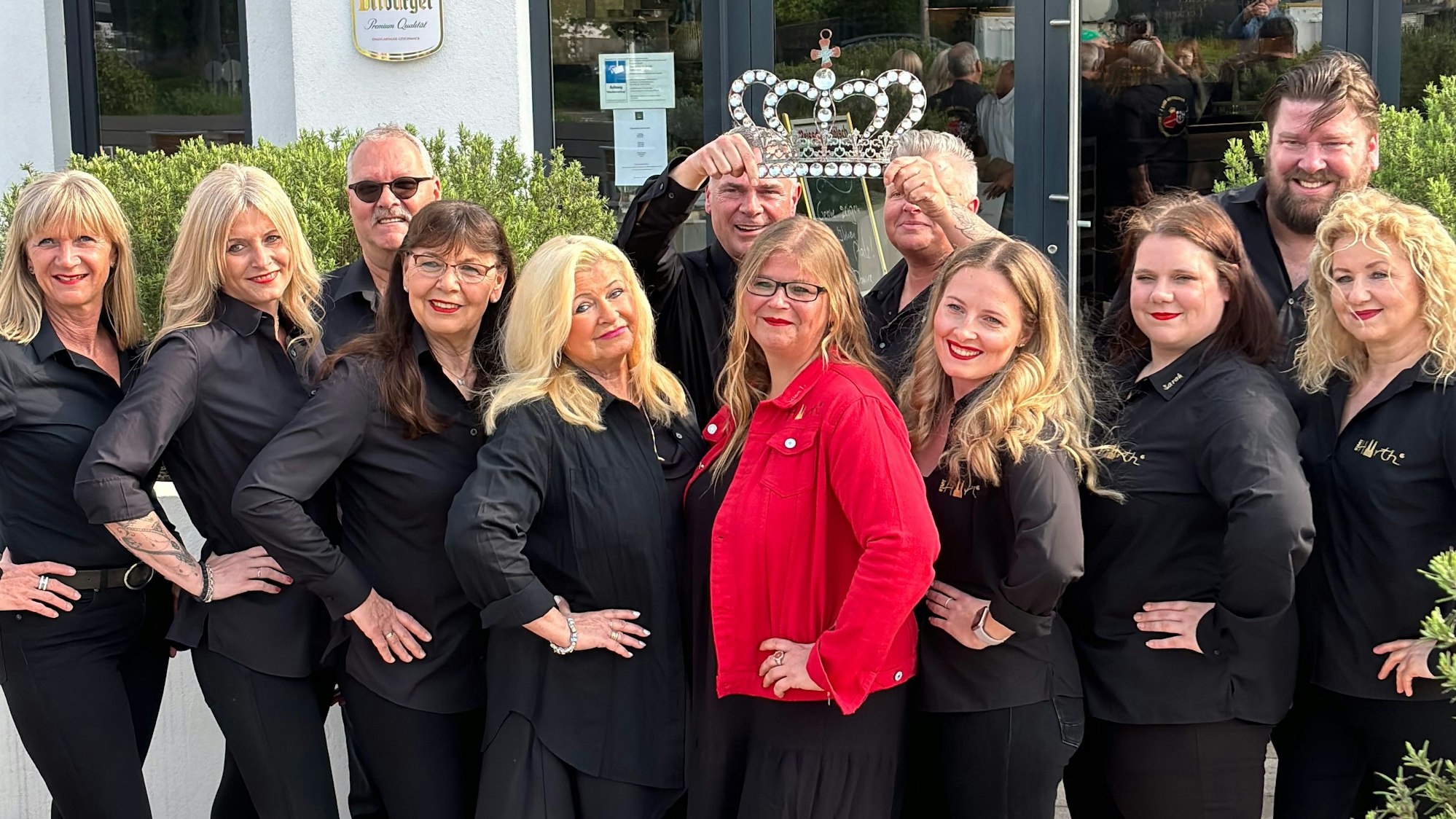 Eine Gruppe von schwarz gewandeten Damen und Herren, in der Mitte die künftige Prinzessin in rotem Blazer und Krone, die von zwei Männern über ihrem Kopf gehalten wird.