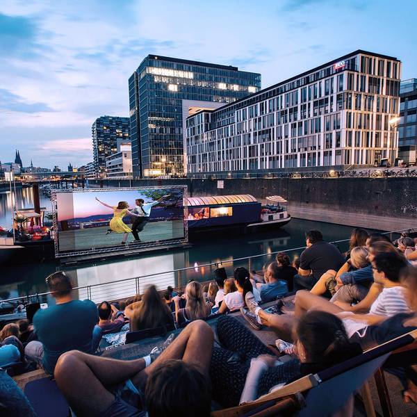 Menschen schauen sich im Open-Air-Kino am Rheinauhafen in Köln vor der Domkulisse einen Film an.