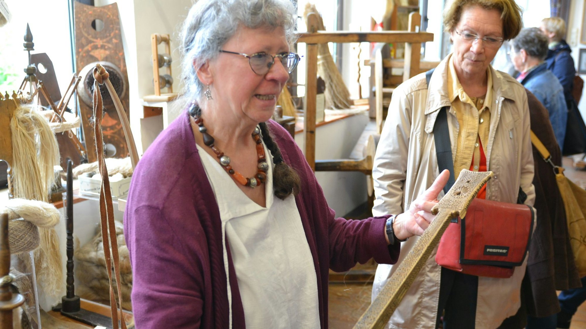 Museumsleiterin Barbara May hält im Handwebmuseum in Bad Münstereifel-Rupperath ein reich verziertes, historisches Werkzeug in der Hand. Eine Besucherin steht daneben und betrachtet es.