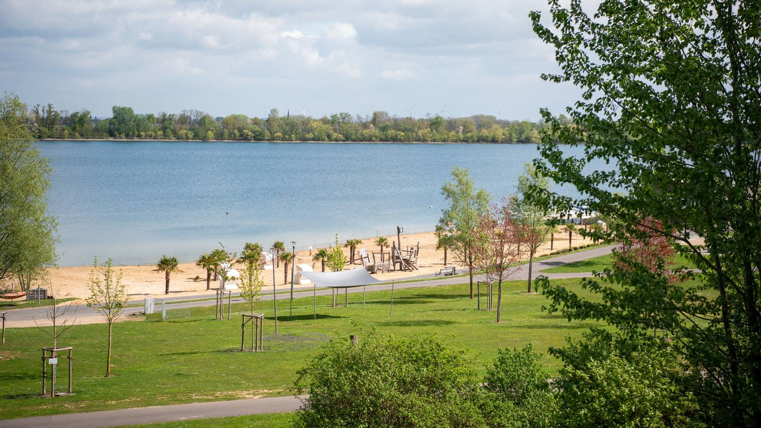 Das Bild zeigt den Wassersportsee in Zülpich mit Sandstrand und Palmen im Frühling.