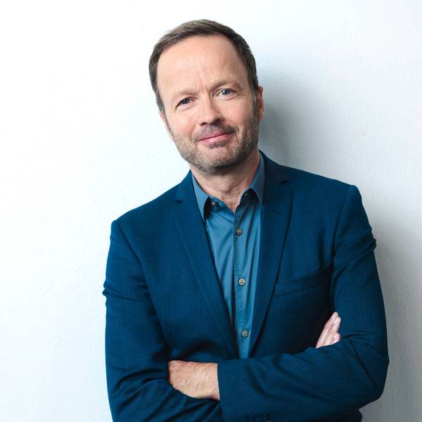 Georg Restle lehnt an einer weißen Wand, er trägt ein blaues Sacko und ein blaues Hemd und lächelt in die Kamera.