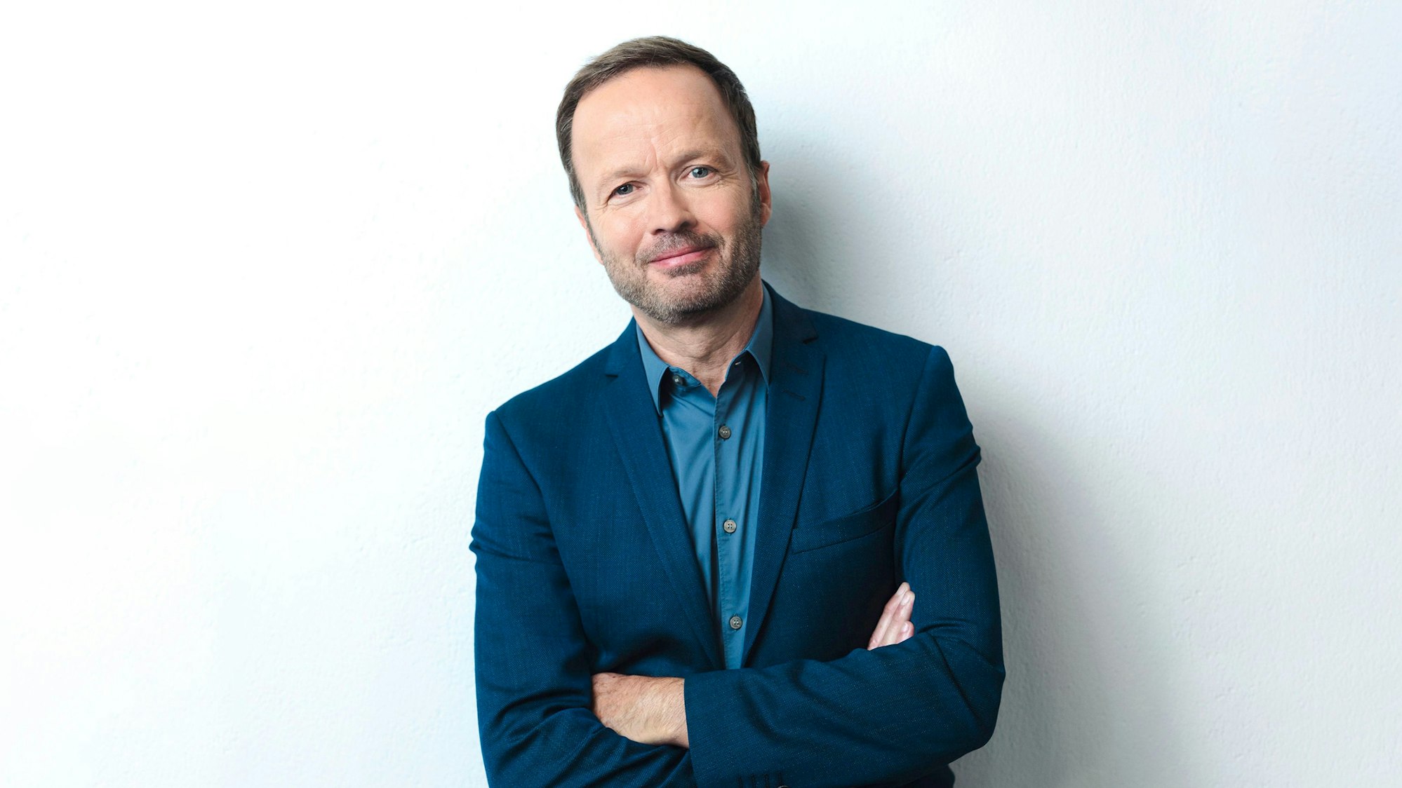 Georg Restle lehnt an einer weißen Wand, er trägt ein blaues Sacko und ein blaues Hemd und lächelt in die Kamera.