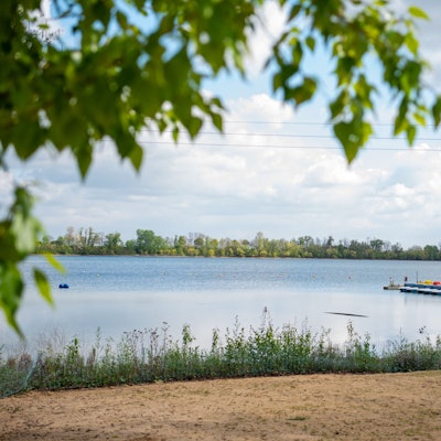 Hinter einem Sandstrand sieht man eine Seefläche. Rechts im Bild ist ein Bootssteg zu erkennen. Um den See herum wachsen Bäume.
