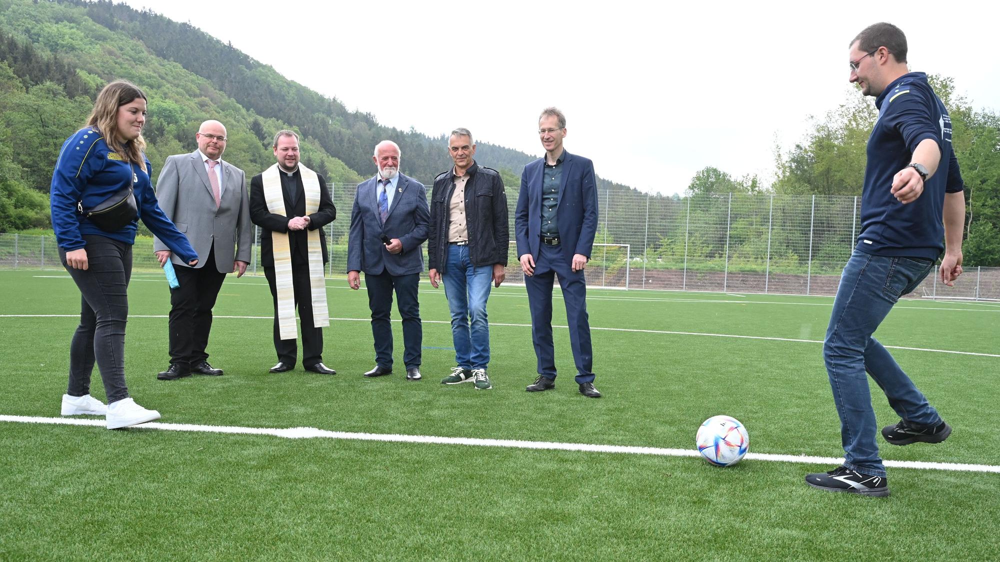 Ein Fußballspieler und eine Fußballspielerin in Jeans und blauen Trainingsjacken spielen sich auf dem neuen Kunstrasenplatz in Hellenthal-Sieberath einen Ball zu. Dahinter stehen fünf Männer und schauen ihnen zu.