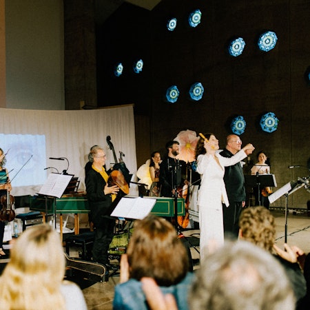 Dorothee Oberlinger steht rechts im Bild vor dem Publikum, die Blockflöte mit der rechten Hand erhoben. Sie trägt weiße Kleidung. Das Publikum ist verschwommen im Vordergrung sichtbar. Im Hintergrund auf der Bühne ist ein kleines Orchester.