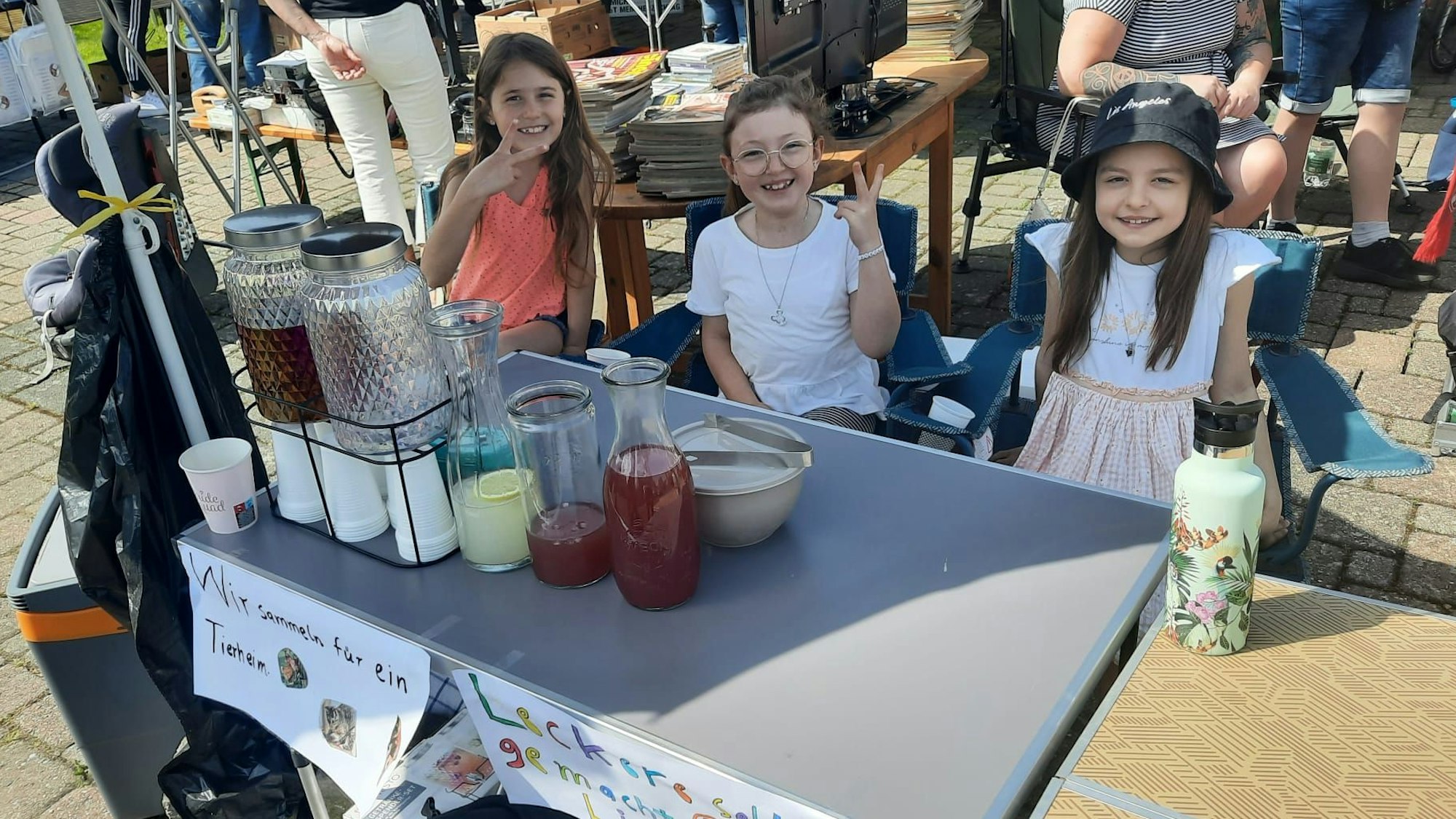 Drei Mädchen sitzen hinter einem Limonadenverkaufsstand.
