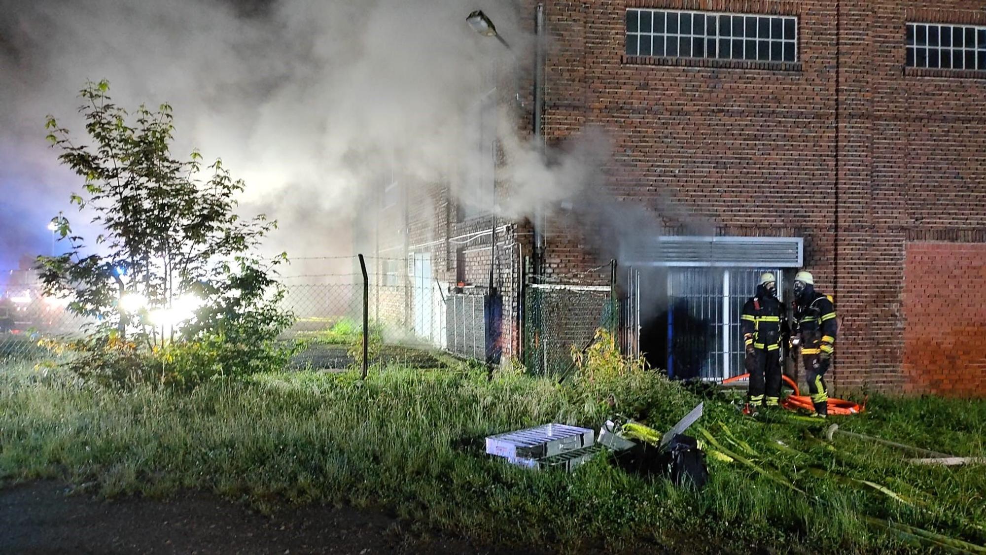 Das Bild zeigt zwei Feuerwehrleute vor der Lagerhalle. Aus den Fenstern dringt Rauch nach außen.