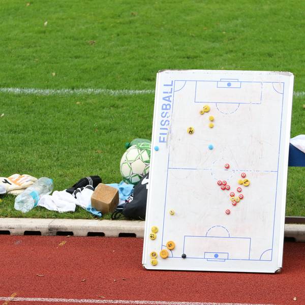 Eine Taktiktafel lehnt an einer Bank, am Rande des Spielfelds liegen Fußballutensilien.