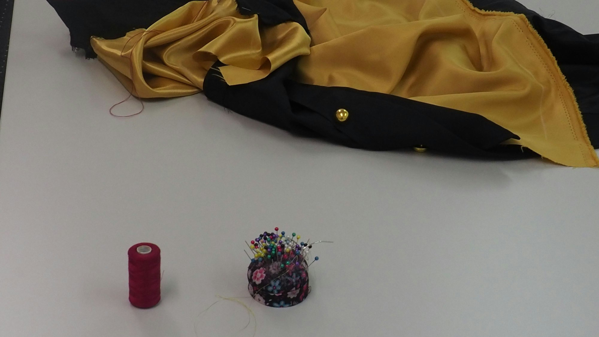 Ein Umhang liegt auf dem Tisch, davor eine Garnrolle und ein Nadelkissen.