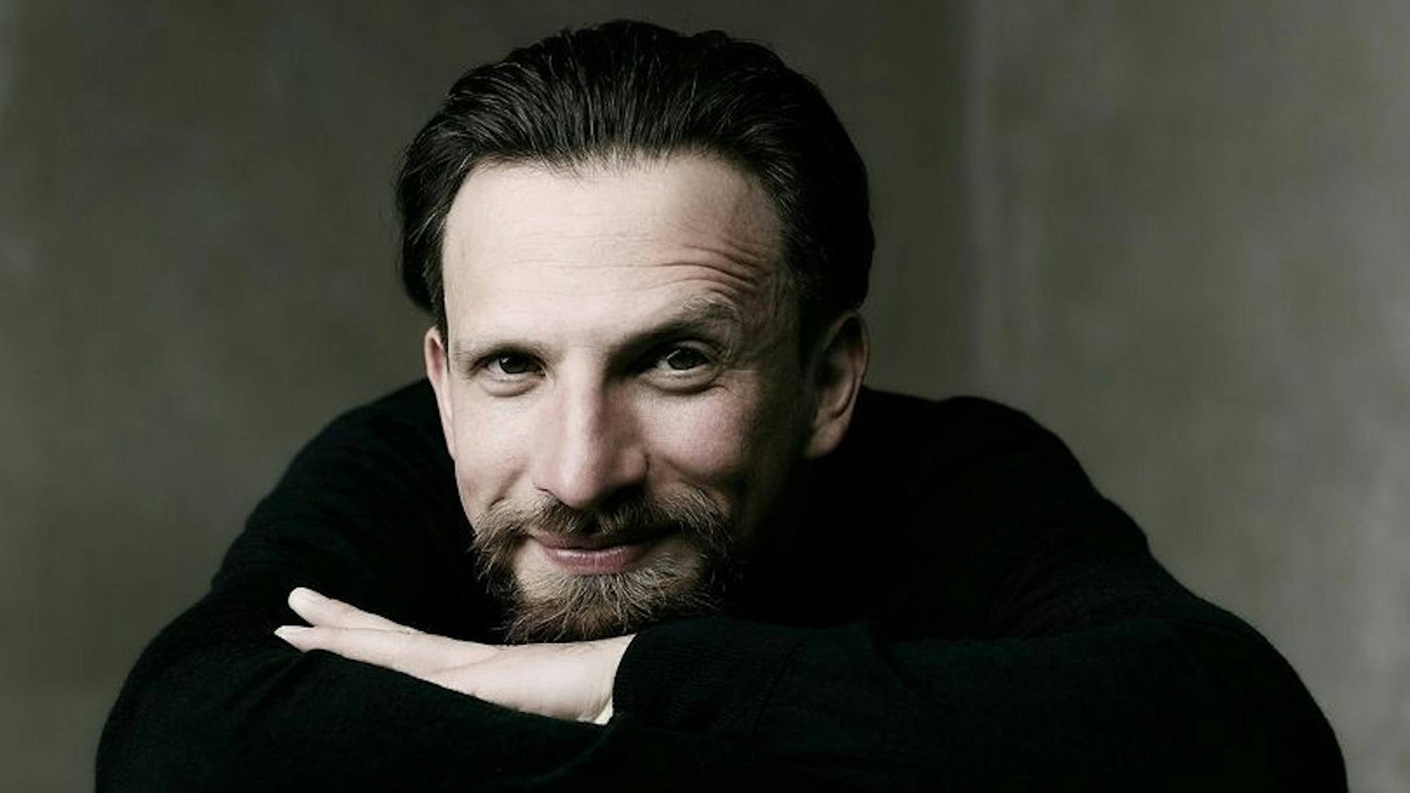 Cornelius Meister hat die Arme überkreuzt und stützt das Kinn auf seine Hand. Er lächelt in die Kamera, hat braune Haare und einen braunen Bart. Er trägt einen dunklen Pullover.