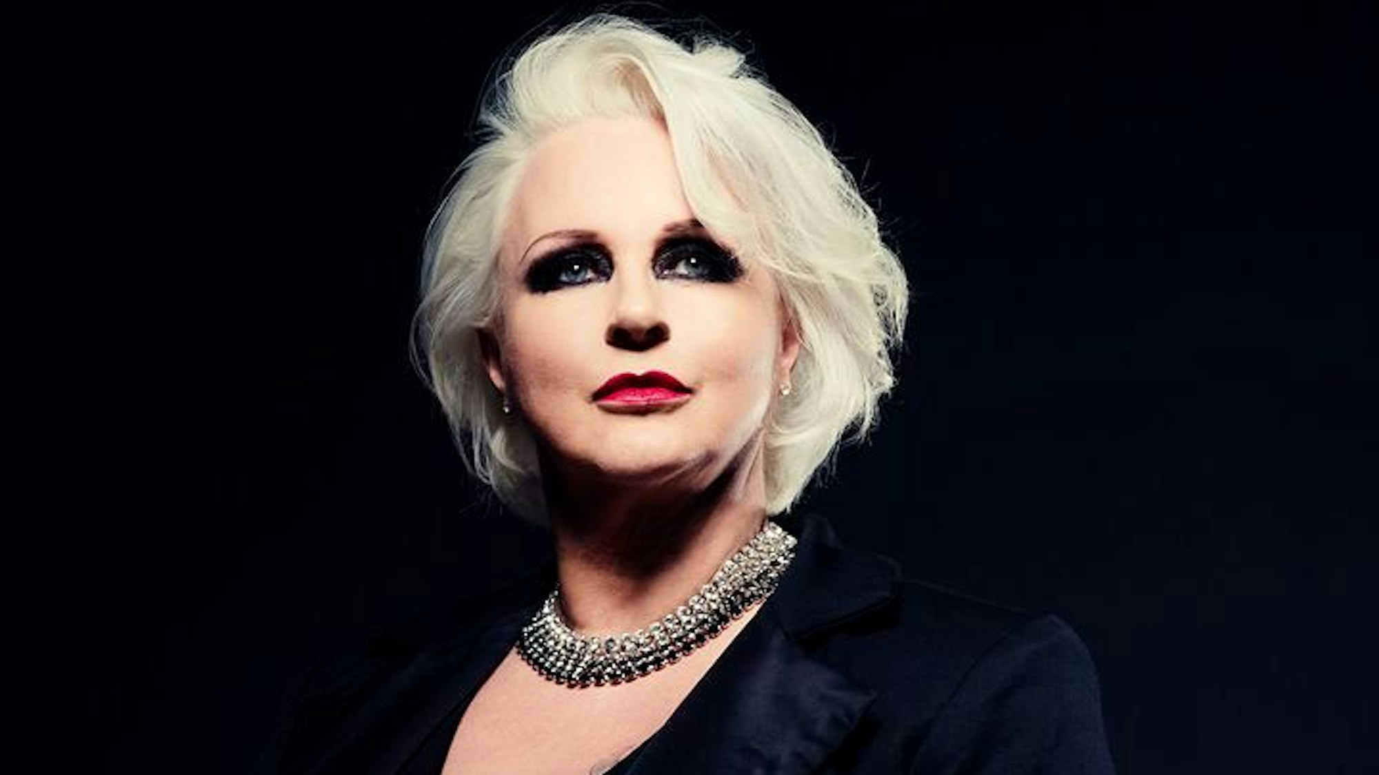 Iréne Theorin hat dunkel geschminkte Augen, weiße Haare und trägt Lippenstift. Sie trägt eine auffällige, glinzernde Halskette und einen schwarzes Sakko. Der Hintergrund ist schwarz.