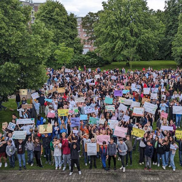 Eine große Menge an Schülerinnen und Schülern steht in der Grünanlage zwischen Bäumen auf dem Schulhof, viele recken Protestplakate in die Höhe.