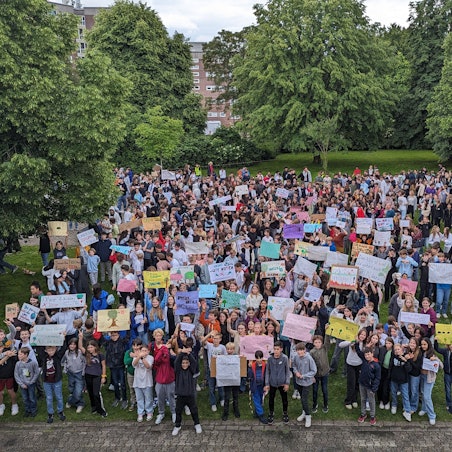 Eine große Menge an Schülerinnen und Schülern steht in der Grünanlage zwischen Bäumen auf dem Schulhof, viele recken Protestplakate in die Höhe.