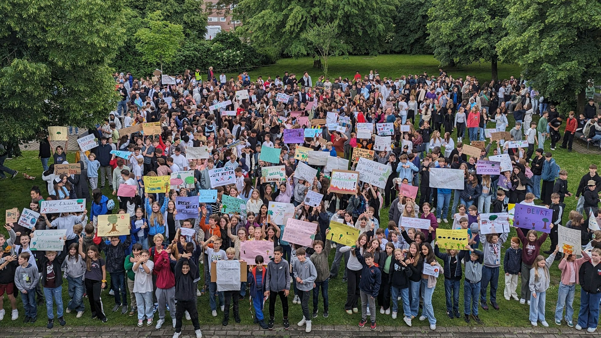 Eine große Menge an Schülerinnen und Schülern steht in der Grünanlage zwischen Bäumen auf dem Schulhof, viele recken Protestplakate in die Höhe.