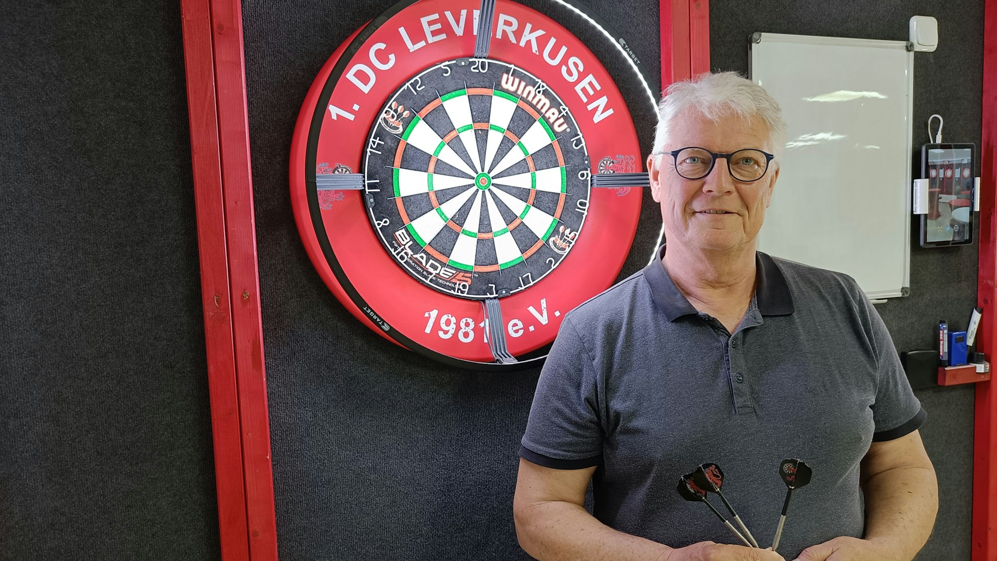 Ein Mann posiert mit Dart-Pfeilen in der Hand vor einer Dartscheibe.