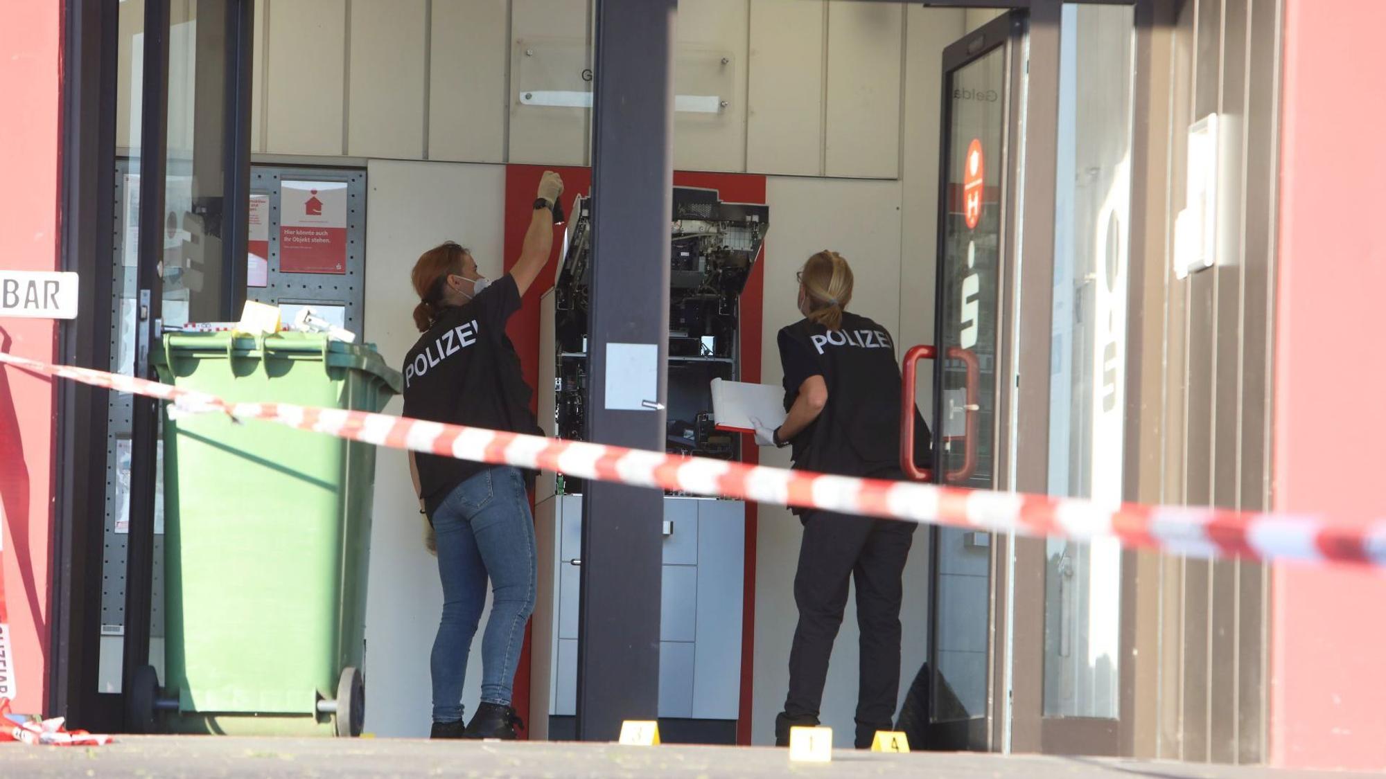 Eine Sparkassenfiliale. Frauen in blauen T-Shirts mit Polizeiaufdruck untersuchen einen zerstörten Geldautomaten.