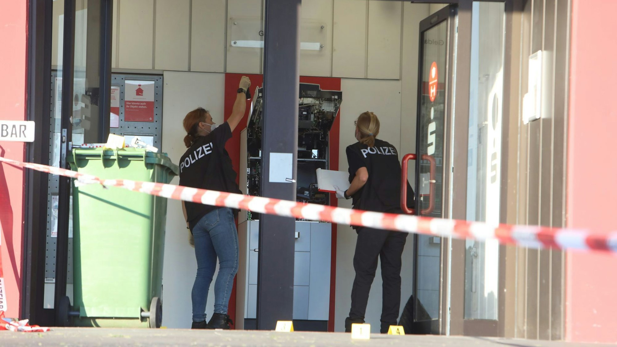 Eine Sparkassenfiliale. Frauen in blauen T-Shirts mit Polizeiaufdruck untersuchen einen zerstörten Geldautomaten.
