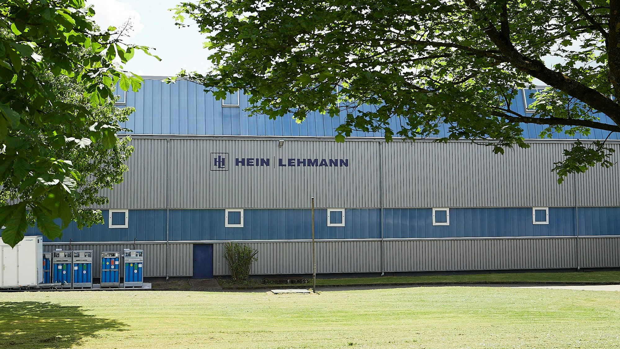 Hinter zwei Bäumen liegt eine der blau-grauen Produktionshallen der Firma Hein, Lehmann in Zingsheim.