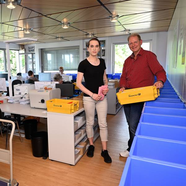 In dem Raum gibt es viele Computer-Arbeitsplätze. Frank Bodengesser hält einen gelben Cotainer mit versandfertigen Briefen in der Hand. Eine Mitarbeiterin zwei Umschläge für die Briefwahl.